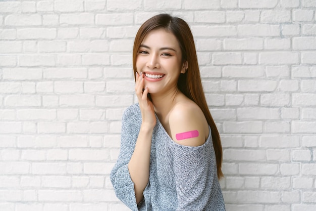 Smiling beautiful asian woman wearing off shoulder sweater\
sleeve showing her arm with adhesive bandage plaster after\
receiving the covid19 vaccine on white brick wall background\
vaccine safe lives