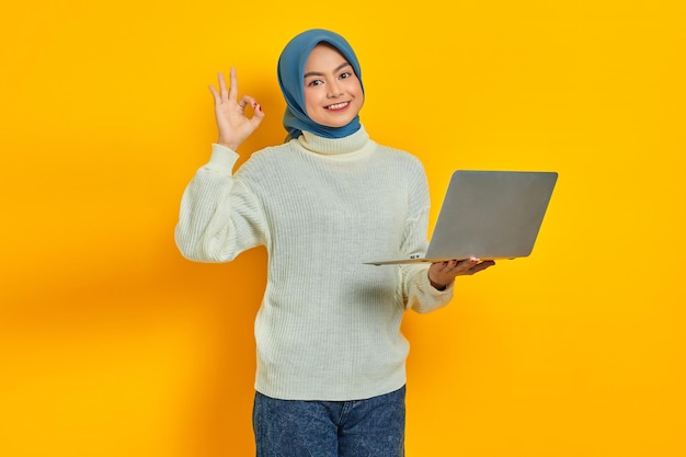 Smiling beautiful Asian woman in white sweater and hijab using laptop and gesturing ok sign isolated over yellow background People religious lifestyle concept