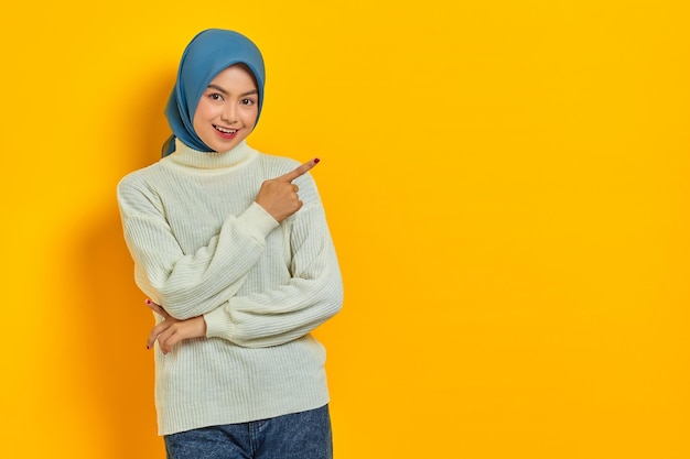 Smiling beautiful Asian woman in white sweater pointing fingers aside at copy space and looking aside isolated over yellow background