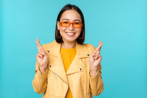 Smiling beautiful asian woman wishing cross fingers for good luck and looking hopeful standing over blue background Copy space