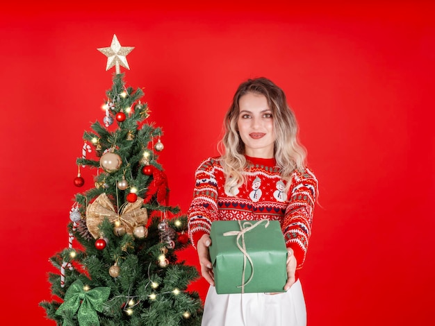 Smiling beautiful blonde caucasian woman giving gift box standing near decorated christmas tree
