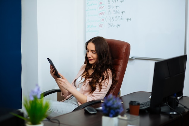 Smiling beautiful business brown-haired lady with long hair sitting at the desk on an office chair looking ahead career growth