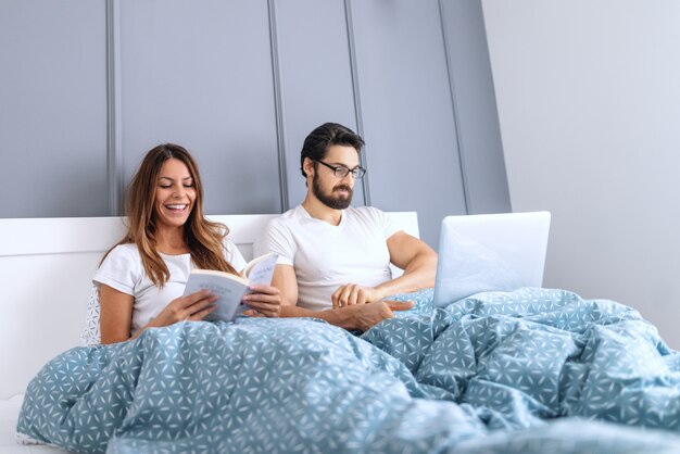 Smiling beautiful Caucasian brunette reading book in bed while her husband with eyeglasses using laptop.