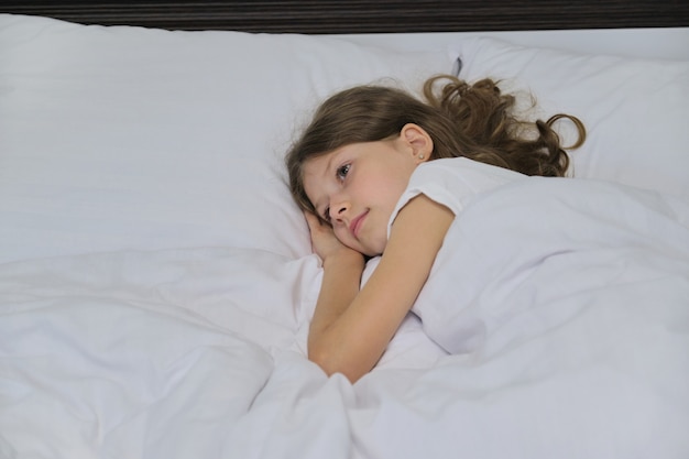 Smiling beautiful child girl lying on a pillow, white bed, close-up face