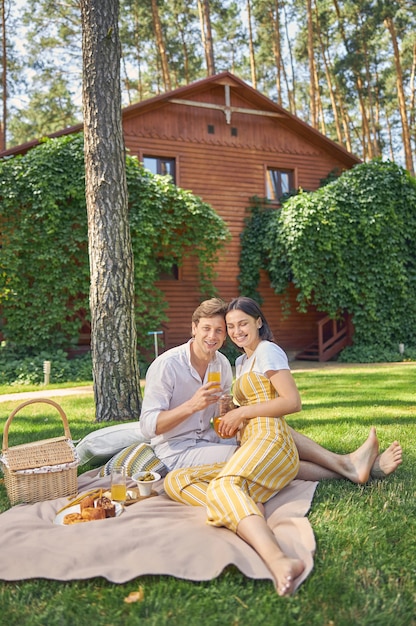Улыбающаяся красивая пара сидит в зеленом саду перед своим деревянным домом