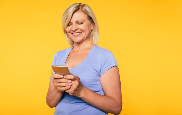 웃고 있는 아름다운 금발의 시니어 여성이 노란색 배경에 격리된 스마트 폰을 사용하고 있습니다