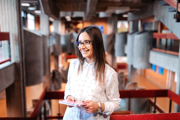 Smiling beautiful female student with brown hair and eyeglasses holding notebook, standing and waiting for class to begin.