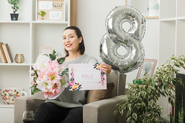 Smiling beautiful girl on happy women day holding bouquet with greeting card sitting on armchair in living room