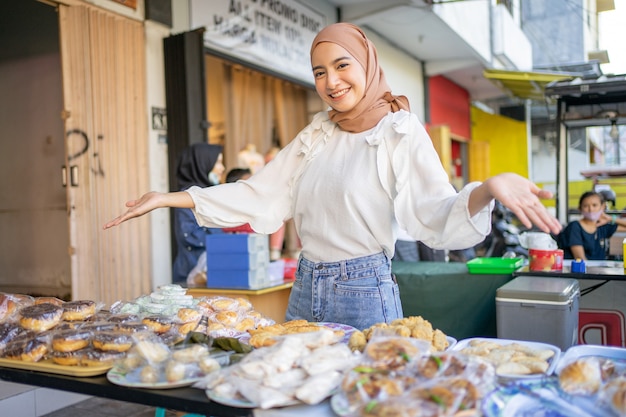 Bella ragazza sorridente in hijab con gesti delle mani che offrono una varietà di cibi fritti