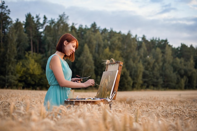 Smiling beautiful girl painter doing her favourite work painting a masterpiece on nature Cheerful girl finishing her painting of golden spikelets