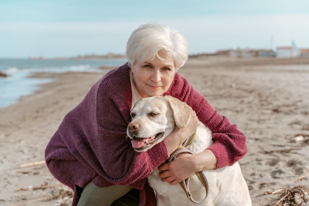 Sorridente bella signora dai capelli grigi seduta sui suoi fianchi che abbraccia il suo simpatico animale domestico sulla spiaggia