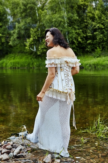 Smiling beautiful mature woman dressed in corset and petticoat is walking along river bank in  wild.