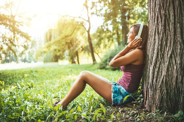 도시 공원의 나무 아래 앉아 스마트폰으로 음악을 들으며 웃고 있는 아름다운 현대 여성