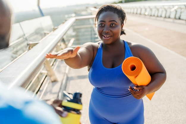 Smiling beautiful plus size african american model holding fitness mat talking with personal trainer