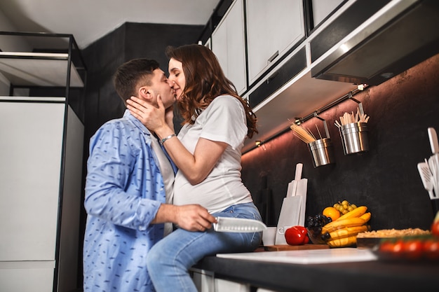 La bei donna ed uomo incinta sorridenti nella cucina bevono il caffè e cucinano. in attesa di una nuova vita, gravidanza.