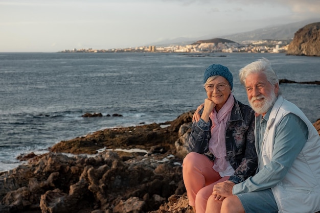 Smiling beautiful senior couple sitting on the rocks at sea\
enjoying travel and nature at sunset light relaxed lifestyle for a\
caucasian couple of retirees