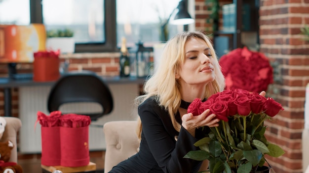 Smiling beautiful woman feeling loved on valentine day smelling elegant bouquet of roses in living room. Affectionate blonde admiring flowers arrangement celebrating anniversary enjoying luxury gift.