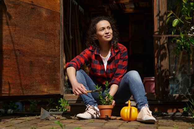 Smiling beautiful woman sitting at the doorstep in a wooden gazebo of a country house and enjoying gardening on a warm sunny day