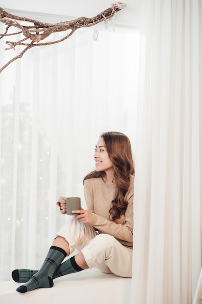 Sorridente bella donna seduta a casa con una tazza di caffè su sfondo di luci.