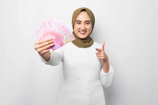 흰색 배경에 고립 된 루피아 지폐에 현금 돈에 hijab 가리키는 손가락을 입고 웃는 아름다운 젊은 아시아 무슬림 여성 사람들의 종교 생활 양식 개념