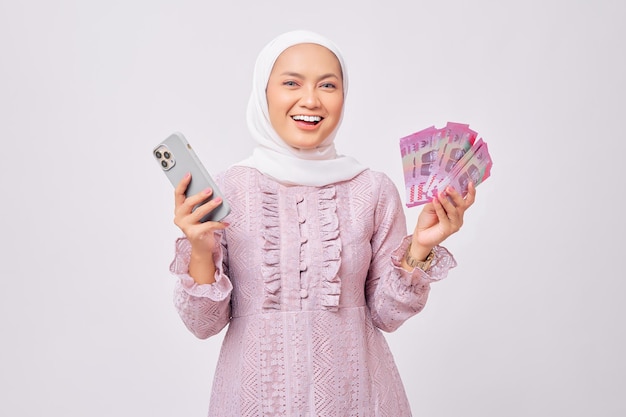 Smiling beautiful young Asian Muslim woman wearing hijab and purple dress holding cash money in rupiah banknote and mobile phone isolated on white studio background