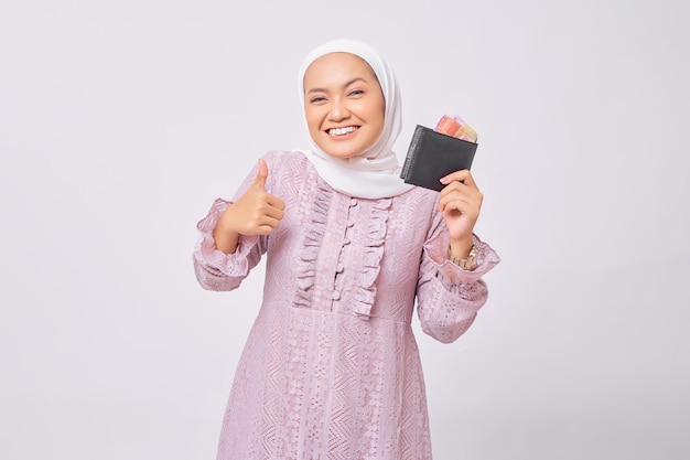 Улыбающаяся красивая молодая азиатская мусульманка в хиджабе и фиолетовом платье, держащая бумажник с полными наличными деньгами и показывающая большой палец вверх жестом на белом фоне студии