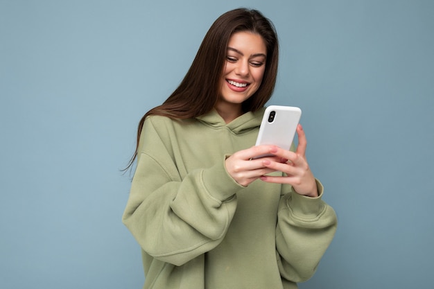 Sorridente bella giovane donna castana che indossa felpa con cappuccio verde alla moda utilizzando il telefono cellulare
