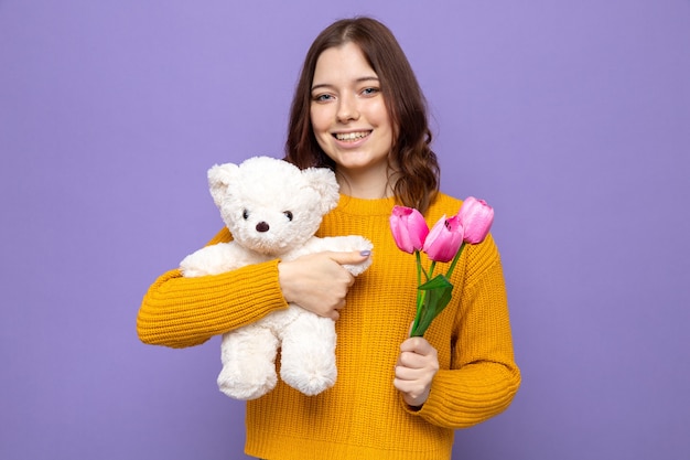 Smiling beautiful young girl holding flowers with teddy bear isolated on blue wall