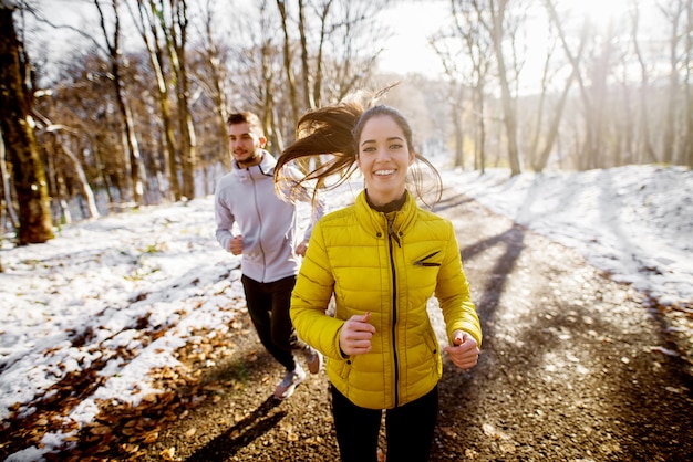 Smiling beautiful young healthy girl running with a trainer in sportswear through the forest in the sunny winter morning.