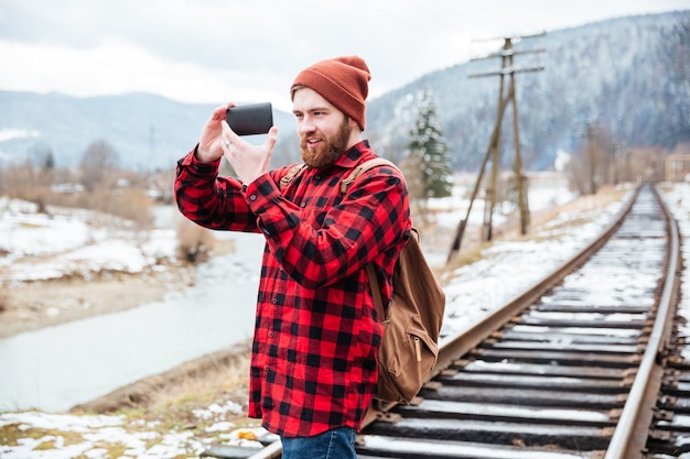 Smiling beautiful young man in checkered shirt and hat taking pictures in mountains