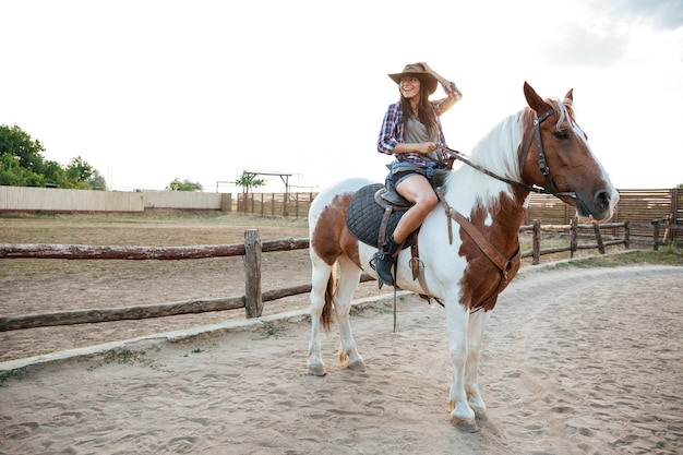 Smiling beautiful young woman cowgirl sitting and riding horse on ranch