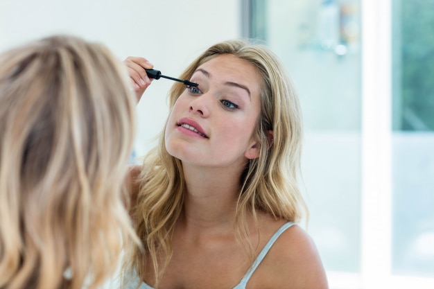 Bella giovane donna sorridente che mette sulla mascara nello specchio del bagno a casa