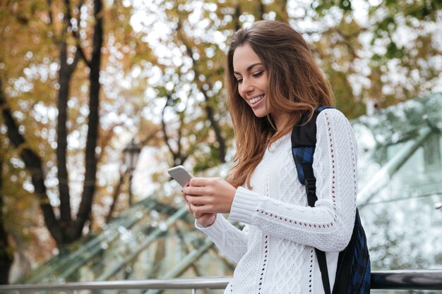 Sorridente bella giovane donna con zaino in piedi e utilizzando il telefono cellulare in park