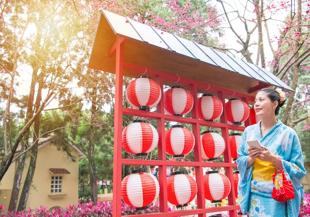 伝統的な着物で夏休みに祭りの情報を検索するモバイル スマートフォンを保持しているお祝いの提灯の装飾の前に立っている笑顔の美しさの女性旅行者。