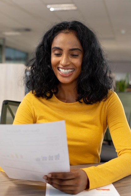 Улыбающаяся двухрасовая деловая женщина анализирует документ, сидя за столом на современном рабочем месте