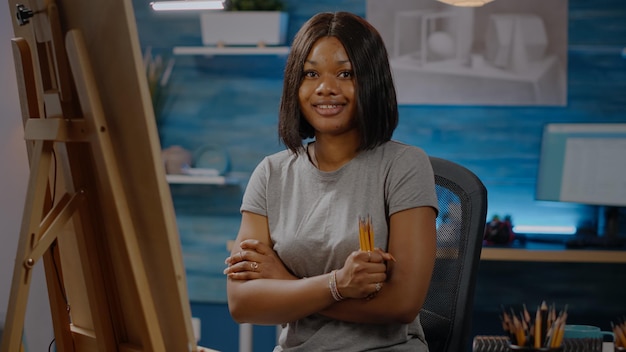 Smiling black artist sitting in art studio space with craft tools and colorful pencils. Woman of african american ethnicity ready for drawing creation on canvas for modern masterpiece