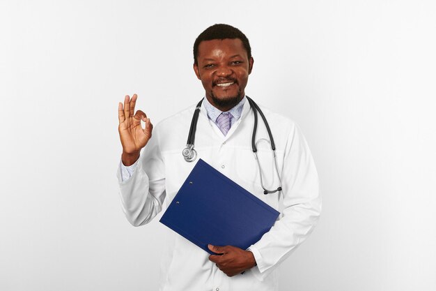 Photo smiling black bearded doctor man in white robe holds medical clipboard and shows ok gesture