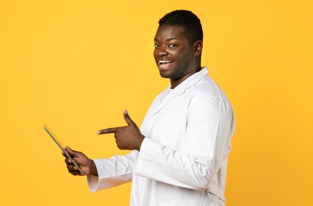 Smiling black doctor pointing at digital tablet on yellow background