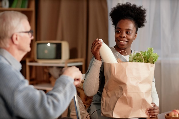 smiling black female caregiver unpacking bag with food delivering groceries to elderly man indoors