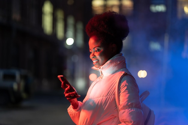 笑顔の黒人の女の子がスマートフォンを持って夜の街で友達を待っている画面読み上げメッセージを見て