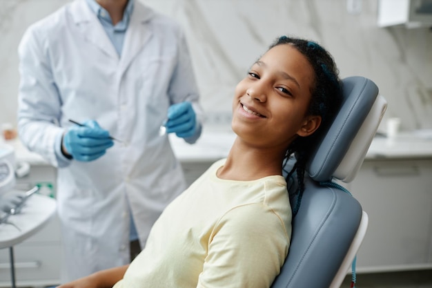 Smiling black girl sitting in dental chair during checkup and looking at camera