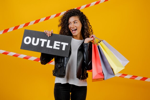 Photo smiling black girl with outlet sign and colorful shopping bags isolated over yellow with signal tape