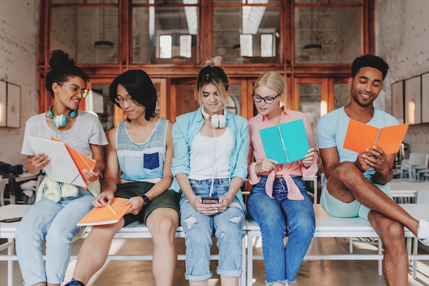 Smiling black guy reading something in orange textbook while his college mates discussing. Pretty curly girl showing asian friend her sketchces in notebook.