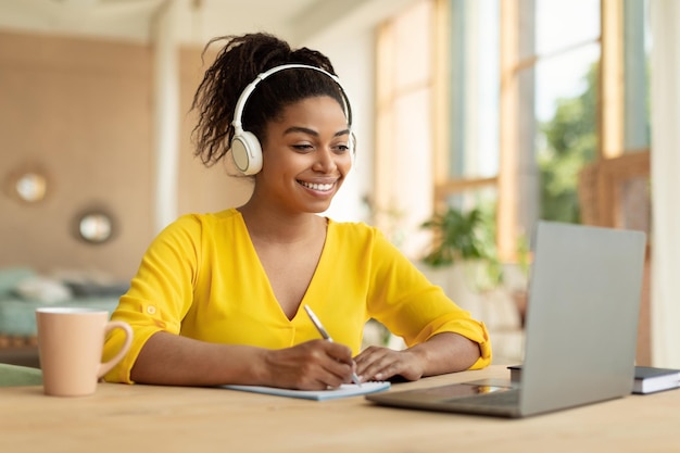 照片微笑的黑人女士在无线耳机在笔记本电脑和笔记在纸笔记本看网络研讨会