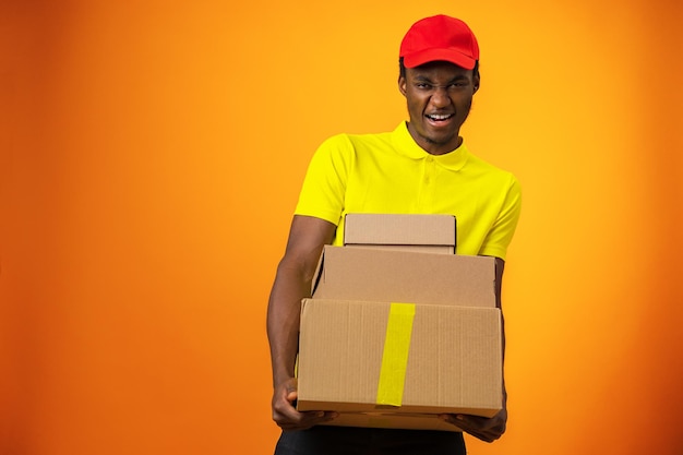 Smiling black male courier wearing uniform holding box in\
orange studio