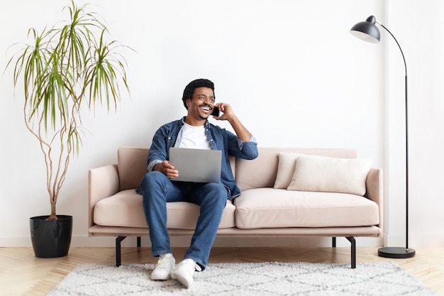 Smiling black man talking on phone and using laptop at home