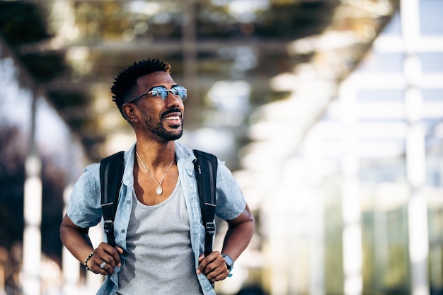 Smiling black man with glasses and backpack walking down the street. Taking the handles of the backpack with his hands.