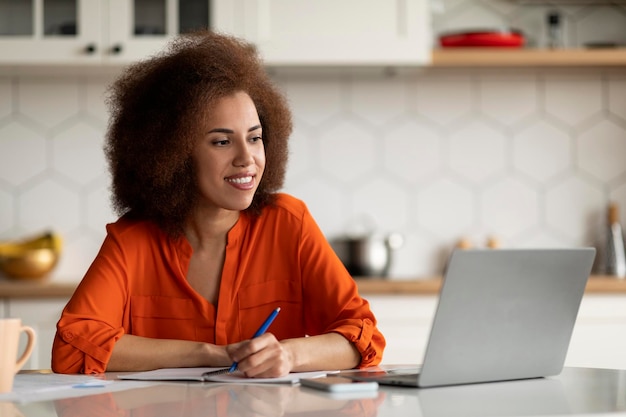 照片微笑的黑人学生女人在厨房使用笔记本电脑和笔记