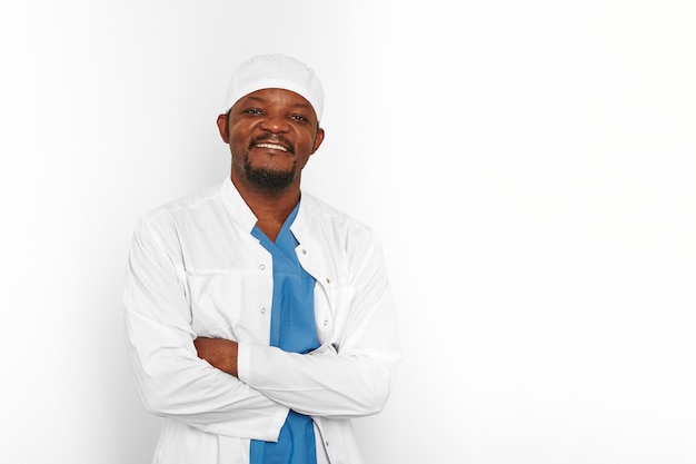 Улыбающийся черный врач-хирург бородатый мужчина в белом халате и кепке со скрещенными руками, изолированными на белом