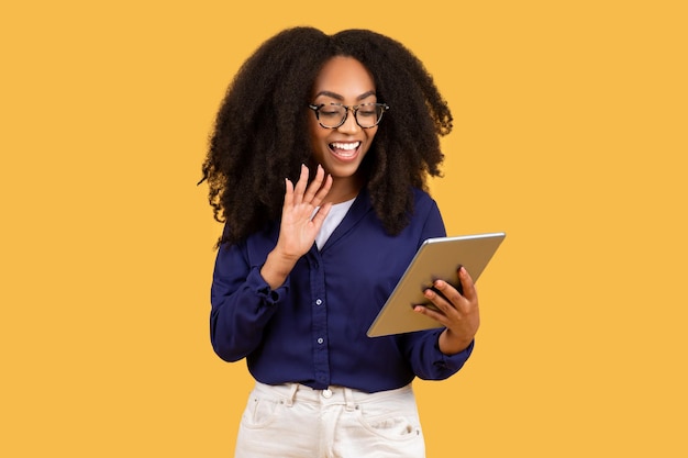 Smiling black woman student use tablet waving hand and having online video call isolated on yellow